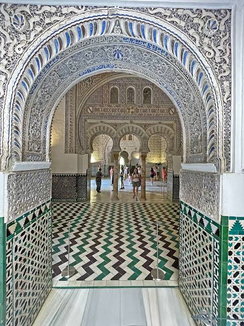 A horseshoe arch doorway in the Real Alcázar of Seville showing the complete Andalusian decorative vocabulary: carved muqarnas arch, zellige tile floor and walls, Arabic inscriptions — the same visual grammar that refugees carried to Morocco in 1492